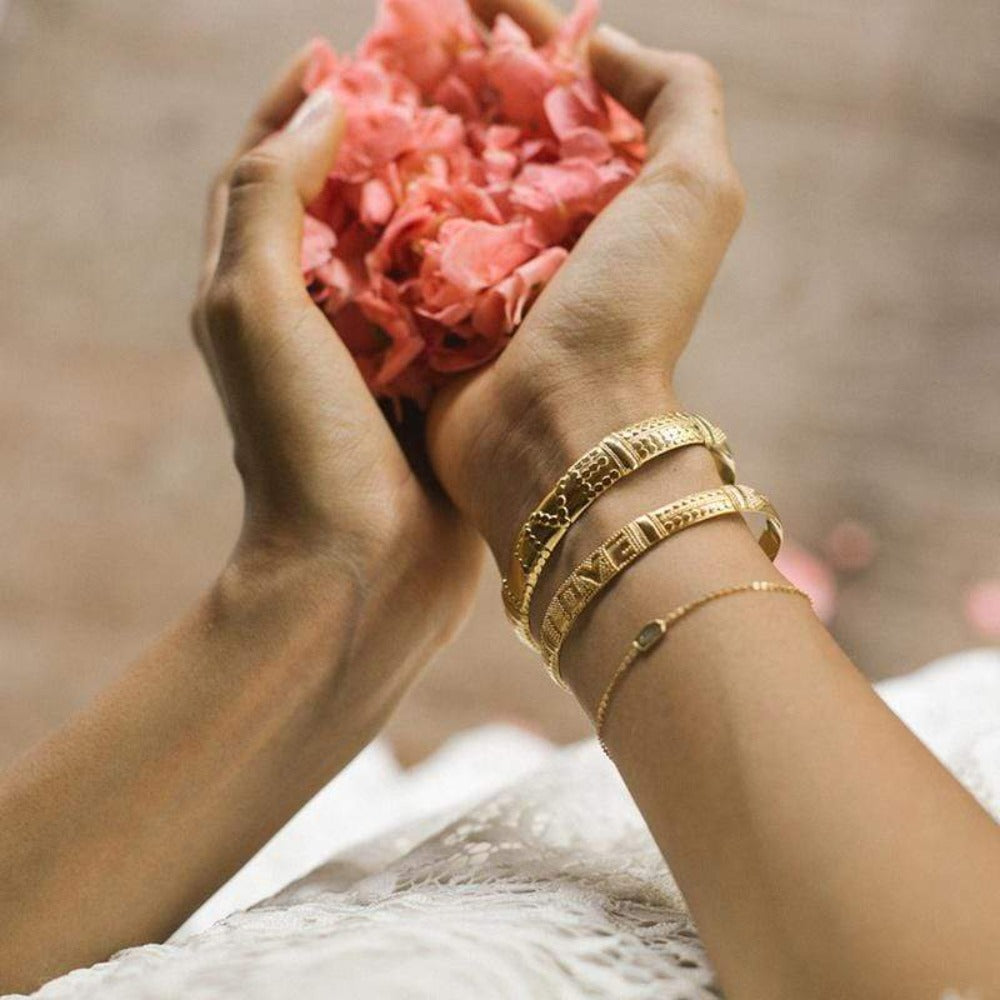 A model holding flower petals and wearing the Love Is Project gold Alchemy LOVE Bracelets